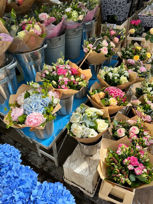 A colorful display of fresh flower bouquets arranged in metal buckets and paper wrapping, featuring pink carnations, white roses, blue hydrangeas, and assorted greenery, placed on a blue table at a fl