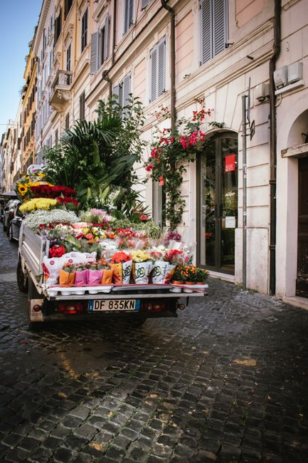 A flower delivery truck parked on a cobblestone street in Charing Cross, with the back loaded with a vibrant and diverse array of fresh floral arrangements. The flowers include bright yellow sunflower