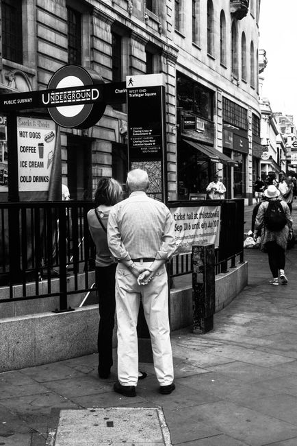 A black-and-white photograph taken in Charing Cross, London, showing two individuals in the foreground—one man with white hair and a woman, both facing away from the camera—standing on the sidewal