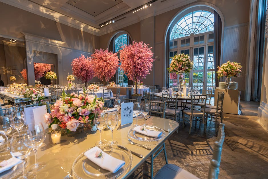 An elegant wedding reception setup within a spacious room featuring large arched windows allowing natural light to illuminate the space. The centerpiece is a large floral arrangement with pastel pink