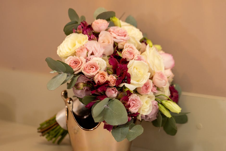 A close-up of a floral arrangement featuring a bouquet of fresh flowers including pale pink and cream roses, small pink spray roses, and deep burgundy blooms, accented with eucalyptus leaves. The bouq
