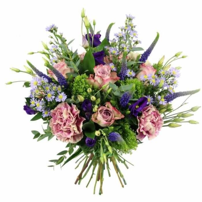 This hand-tied floral arrangement, photographed against a pure white background, showcases a sophisticated blend of pinks, purples and greens that would look perfectly at home in a stylish Charing Cross townhouse or a chic office near Embankment. The bouquet is composed in a rounded, slightly dome-like shape, with its focal point a collection of soft pink roses at different stages of opening. Some blooms remain in tight, elegant buds, their petals wrapped close and tinged at the edges with a slightly deeper pink, while others reveal more of their spiralled centres, the colour softly fading toward cream at the outermost petals. Tucked among the roses are generous carnations in muted mauve and dusty rose, their lavishly ruffled petals lending a full, cushiony texture that fills out the arrangement. Standing proud above the rounded blooms, tall spires of deep violet-blue Veronica introduce slender, architectural lines, giving the bouquet height and a sense of movement that echoes the rhythm of pedestrians flowing through Charing Cross. Between these vertical elements and the rounded blossoms, rich purple lisianthus flowers, some open and others still in bud, add velvety intensity, balancing the pastel tones. Sprinkled throughout, small, aster-like blossoms in soft lavender and periwinkle form airy sprays of colour, their star-shaped petals and pale centres creating a delicate, almost cloud-like veil. The flowers are supported by abundant foliage: bright lime-green textural filler flashes vibrantly against more subdued eucalyptus leaves, whose cool, silvery hue and rounded shapes lend a soothing backdrop. Additional greenery in varied tones completes the lush, natural look. At the base, the stems are clearly visible, neatly tied and trimmed, ready to be slipped into a favourite vase. The bouquet projects an air of polished elegance with a romantic twist, ideal for birthdays, anniversaries, or thoughtful thank-you gestures sent within the Charing Cross area.