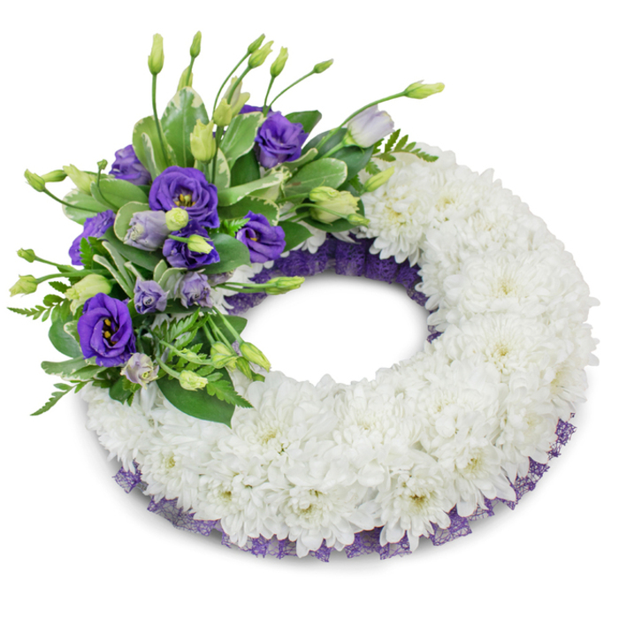 This elegant circular funeral wreath is shown in a studio setting against a stark white backdrop, every detail arranged to convey quiet respect and remembrance. The dominant body of the wreath is formed from countless white cushion chrysanthemums, densely packed so that their ruffled petals create a uniform, velvety ring - a tactile, peaceful field that symbolises purity and enduring affection. Positioned deliberately in the upper-left sector, a rich cluster of purple Eustoma (lisianthus) provides the single point of colour: fully opened deep-violet blooms sit alongside softer lavender buds, their layered petals and subtle tonal shifts producing depth and a comforting visual rhythm. Fresh green foliage - glossy, variegated leaves and feathery fern-like sprays - nestles among the lisianthus stems, offering texture and a gentle, living contrast to the pristine white base. A delicate trimming of dark purple, mesh-like ribbon runs just beneath the flowers along both inner and outer edges, its crinkled lace effect harmonising with the lisianthus hue and giving a refined border to the composition. Soft, diffused lighting and the faint shadow beneath the wreath emphasise its roundness and the craftsmanship of the construction. As a sympathetic tribute suitable for memorial services across Charing Cross, close to Northumberland Avenue and the theatres on the Strand, this wreath is designed to read as dignified and consoling - a carefully balanced expression of sympathy and remembrance.