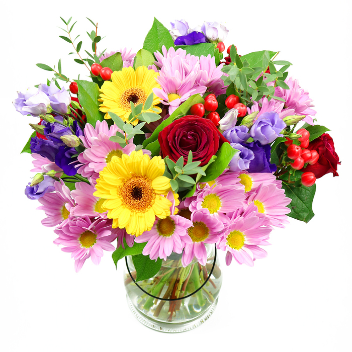 A beautifully balanced mixed bouquet stands tall in a clear glass vase, centred against a pristine white backdrop that highlights every subtle detail. At the core of the arrangement, rich deep red roses unfurl in velvet-like layers, lending the bouquet a sense of depth and heartfelt sentiment. Flanking these roses, two bright yellow gerbera daisies, each with a dark brown-orange centre, project forward like warm smiles, adding a joyful, sunlit quality that feels perfectly in tune with the lively energy of Charing Cross and the bustle of nearby Trafalgar Square. Encircling these hero blooms, generous clusters of soft pink daisies or chrysanthemums create a halo of pastel softness; their myriad slender petals and small yellow hearts build a feathery texture and a feeling of abundance. Interspersed among the pink daisies are elegant lisianthus blossoms in shades of purple and lavender, their gently fluted, bell-shaped flowers introducing cool tones and a sophisticated, slightly whimsical air. Throughout the bouquet, glossy red hypericum berries appear in neat clusters, their smooth, rounded forms offering a distinct contrast to the open petals and infusing a contemporary edge. The flowers are embraced by layered greenery that ranges from dark, broad leaves to fine, light-green shoots that arc outward, softening the silhouette and hinting at a natural, garden-fresh origin. The clear vase reveals the trimmed stems and clear water below, grounding the airy composition and showcasing the florist's precise handiwork. The result is a bouquet that feels both polished and inviting, ideal for same-day delivery to a townhouse just off the Strand, a hotel lobby near Charing Cross station, or a private celebration overlooking the river.