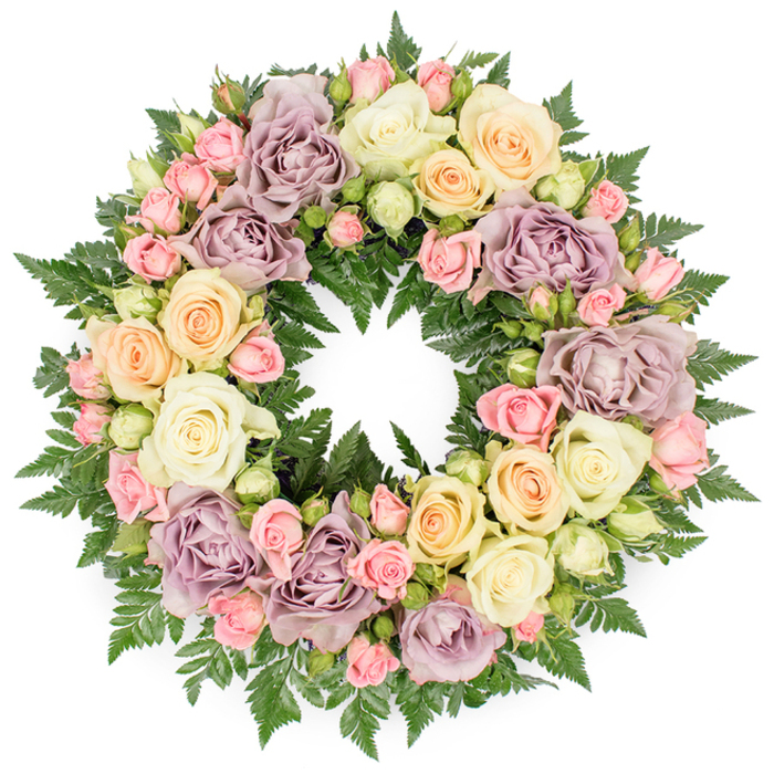A lush, circular funeral wreath filled entirely with pastel roses and deep green foliage is presented on a clean white backdrop, highlighting its form and intricate craftsmanship. The wreath's ring is complete and even, with blossoms arranged so closely together that they overlook one another, creating a layered effect that seems to draw the viewer inward. Large, fully open roses in creamy ivory and soft white form the main structure, their velvety petals curled loosely at the edges, giving a sense of softness and ease. Interspersed among them are delicate blush pink spray roses, modest pale peach blooms, and touches of buttery yellow that infuse the design with a gentle warmth. Distinctive dusty lavender or muted mauve roses add a subtle, heirloom quality, reminiscent of vintage floral prints and family keepsakes often treasured in homes around Charing Cross and the nearby West End. Between the flower heads, fern fronds and fine greenery weave through the circle, their rich dark green tones deepening the contrast and giving the pastel palette something solid to rest against. The textures shift from the smooth, satin-like rose petals to the slightly rough, feathery surfaces of the ferns, creating visual interest and movement. The soft, steady light in the image gives the wreath a calm presence, like a quiet pause amid the energy of central London. This arrangement, expertly styled in the tradition of local funeral floristry, would sit comfortably at a service, wake, or memorial reception near Charing Cross, offering a tender expression of sympathy and a graceful symbol of everlasting remembrance.