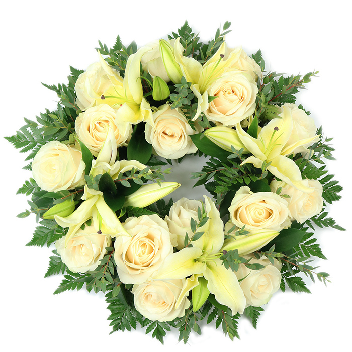 A carefully composed, circular funeral wreath sits photographed from above on a white field, its palette restrained to creams, pale yellows and varied greens. The design centres on roughly fifteen ivory roses, each bloom with classic, tightly folded petals that reflect light softly and create a unified, luminous field. Between the roses, pale lemon lilies open in elegant, trumpet-shaped forms-some fully revealed with graceful petals and subtle stamens, others held as poised buds that introduce vertical contrast and promise. Supporting the blooms is an abundant bed of greenery: feathery fern fronds produce a delicate, lacy texture around the wreath's perimeter while clusters of rounded, darker foliage-likely eucalyptus or a similar leaf-give a matte, blue-green counterpoint and broaden the composition's tonal range. The wreath's dense construction produces a full, comforting visual weight yet retains a measured openness at the flower margins so the eye can rest on individual blooms. Lightly scented, the piece suggests the soft sweetness of roses with a crisp, green edge from the lilies-an aroma suitable for intimate interiors such as a chapel on Villiers Street or a quiet funeral parlour near Whitehall. The circular form is deliberate, symbolising memory and continuity; the colour scheme of cream, lemon and green underscores purity, hope and solace. Prepared with experienced hands and a respect for traditional sympathy floristry, this wreath is intended as a dignified memorial tribute or graveside offering for families in and around Charing Cross and the Northbank.