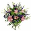Set against a clean white backdrop, this hand-tied bouquet feels like a moment of calm luxury discovered just off the Strand, its rounded form and layered colours drawing the eye in the way evening lights draw people toward Charing Cross. At the centre are soft, romantic pink roses, some still cupped tightly with petals edged in a deeper blush, others beginning to open into gentle swirls that drift towards pale cream at the tips. Nestled beside them are lush carnations in dusty rose and mauve, their many ruffled petals forming plush clusters that bring a sense of depth and comfort, like a soft fabric gathered in the hand. Pushing gracefully through this sea of petals, tall stems of deep violet Veronica stand like elegant markers, their slender, tapering blooms adding height and a slightly modern edge. Rich purple lisianthus blooms, along with their smooth, pointed buds, are dotted throughout, offering velvety shadows that make the surrounding pastels appear even more luminous. Light sprays of delicate aster-like flowers in lavender and periwinkle weave between the larger blossoms, their tiny star-shaped petals and soft centres giving the whole arrangement a gentle, airy halo. Around the flowers, greens in multiple tones keep the design fresh: vivid lime-green textural filler glows from within, while muted, silvery eucalyptus leaves and other foliage provide a cool, calming frame. The cleanly cut stems gathered at the base underscore its hand-crafted nature and make it easy to place straight into a vase on a desk near Trafalgar Square or a windowsill looking toward the river. The overall mood is one of quiet celebration and affection, perfectly suited for birthdays, heartfelt thanks, or simply sending a beautiful burst of colour to someone in the Charing Cross neighbourhood.